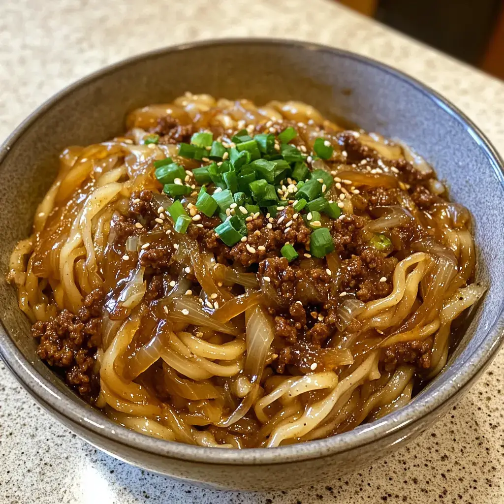 A bowl of Mongolian ground beef noodles with caramelized onions, scallions, and sesame seeds, photographed from above for a rich, savory presentation.

